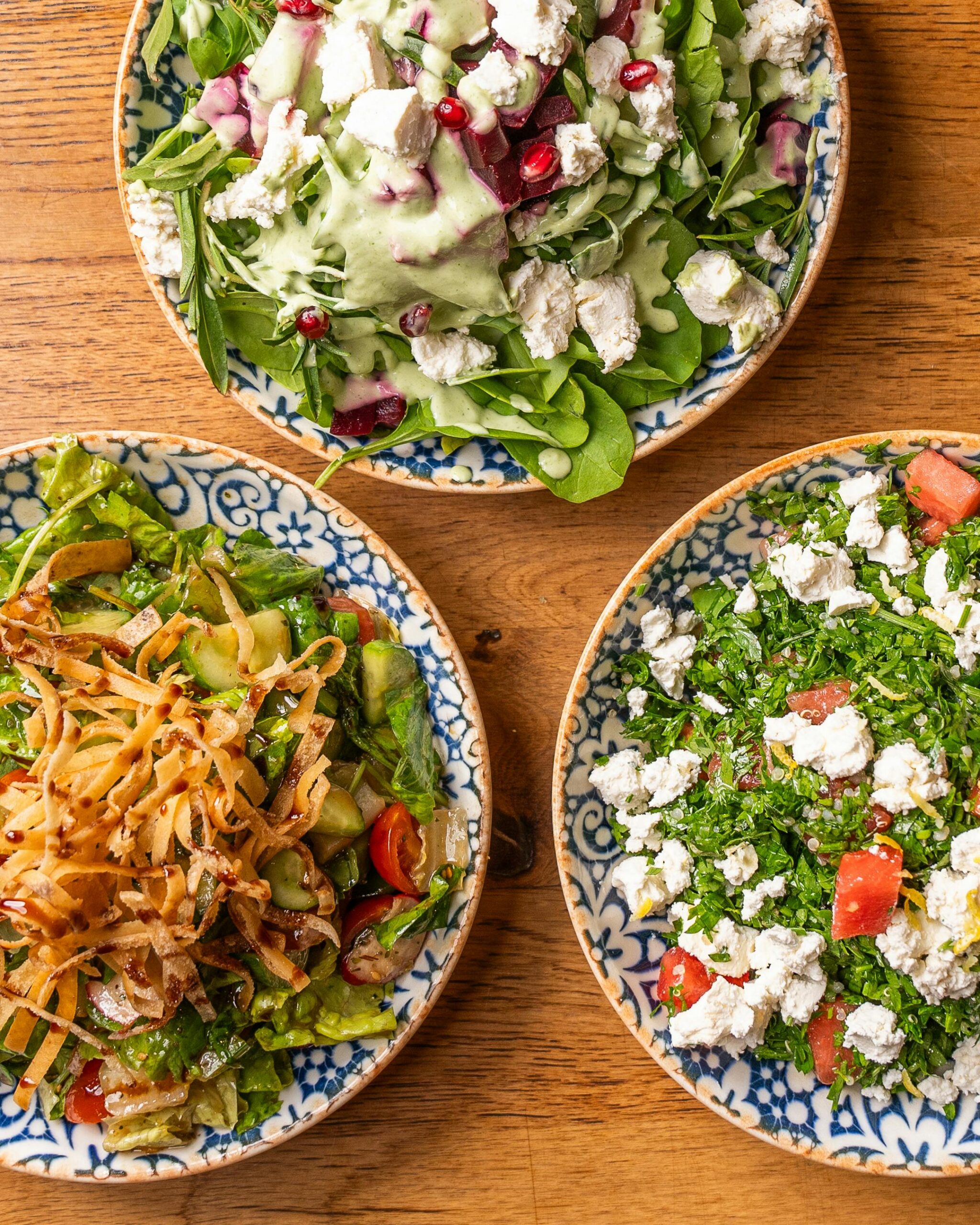 Top view of three fresh salads with feta cheese, vegetables, and herbs on a wooden table.